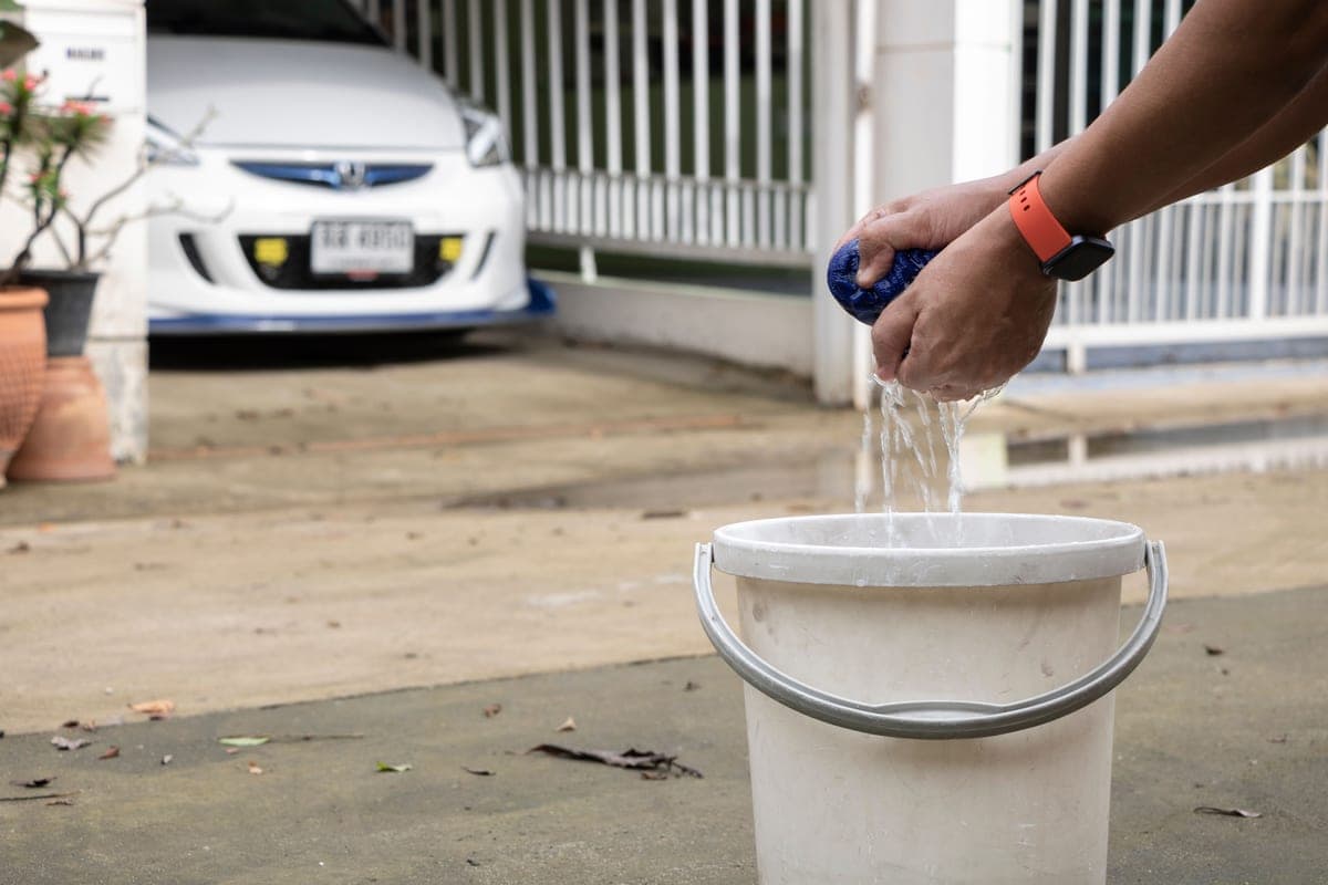 Two wash buckets and a microfiber mitt beside a car being hand-washed